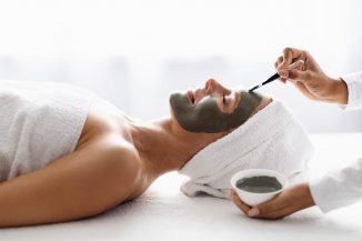 Cosmetologist making skincare beauty treatments to smiling middle aged woman in luxury spa salon, applying moisturising clay mask on face of relaxed mature female, side view shot with free space